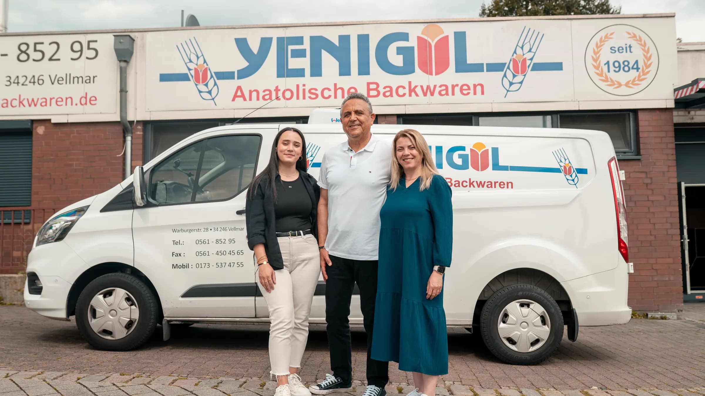 Familie Yenigül, zwei Frauen und Mustafa Yenigül, vor einem Lieferwagen und dem Firmenschild mit dem Siegel „seit 1984“